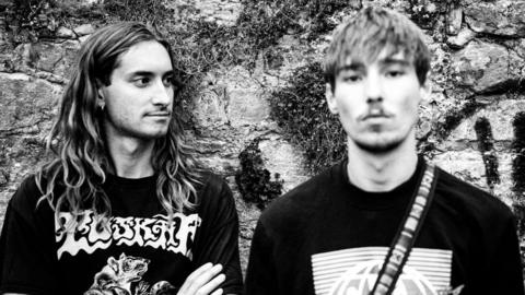 Two young men, one on the left with long hair and one on the right with shorter hair and a light beard, shot in black and white