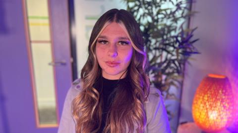 A woman with brunette hair and blonde highlights wearing a grey jacket. She is in a room with blue lighting. There is a lamp and plant in the background.