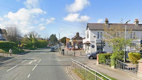 A road with metal fences on either side and some shops and a few houses. There is a petrol station in the distance and some cars on the road.