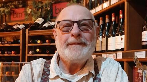 Ken Langley has short grey hair and a grey beard. He is wearing dark framed glasses and a white t-shirt, white shirt and paisley patterned braces. He is holding a wine glass and is sat in front of shelves full of wine bottles.
