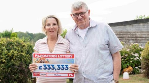 Rebecca Andrews and her husband David Docherty hold a large check with £333,333 on it. They are standing on a grassed lawn surrounded by shrubs and bushes.