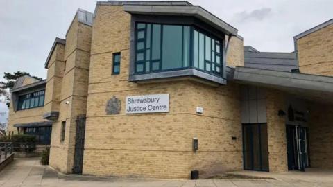 The entrance to a pale brown brick building with dark windows and the words "Shrewsbury Justice Centre" on the side.