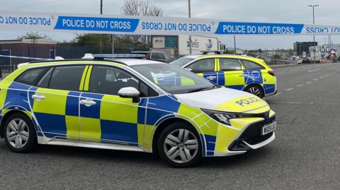 Two police cars with blue and white livery are parked close to a line of blue and white police tape on a road