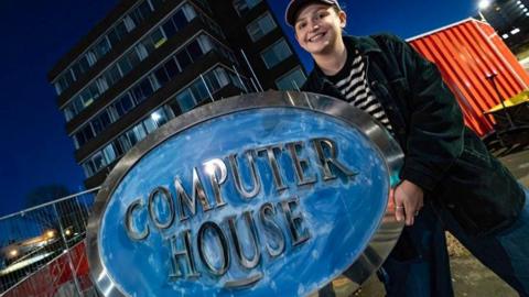 Edie Miller holding the large blue Computer House sign. She is standing in front of the building which is being demolished.