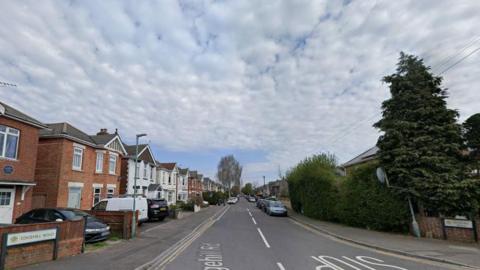 Road with houses stretching down on both sides and cars parked on drives and on the road.