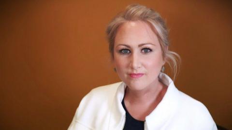 Llinos Griffin-Williams standing in front of an orange background. She is looking at the camera. She has her hair tied back and is wearing a white jacket.