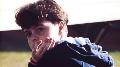 Picture of Emma-Jayne Knowles looking over her shoulder towards the camera with her hand in front of her face. She has brown wavy hair and is wearing a blue jacket.