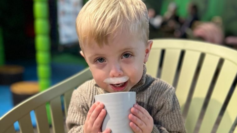 Hudson is wearing a brown jumper and drinking from a cup and has what looks like milk left on his top lip. He has blond hair and is sitting in a light green chair
