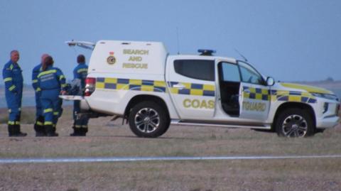 A white Coastguard vehicle with the words Search and Rescue and Coastguard written on the site. It is on a beach and beyond it is sea. There are four people in blue boiler suits at its rear door which is open.