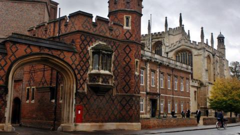 Eton College. A photograph of the redbrick school taken from across the road. A large archway is shown leading into the building on the right. Smartly dressed people are standing in a yard outside the adjacent building.