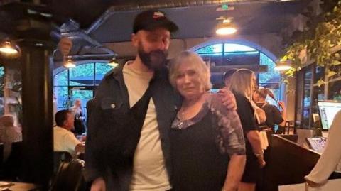 Philippa, wearing black dress, and Joe, wearing baseball cap, stood in restaurant hugging.