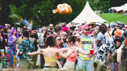 A group of festival-goers in brightly-coloured clothing looking happy and relaxed. Two are dancing in the foreground and in the background there is a large tipi tent.