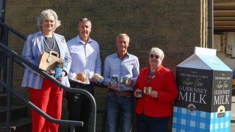 Four people stood on steps. On the left is a woman wearing red trousers, a black top, striped blazer and has short blonde hair. She is holding a brown paper back and box of snacks. On the right of her is a man in a light blue top and he is holding scones. To the right of him is a man wearing a light blue top, blue jeans and holding Guernsey Dairy products. To the right of him is a woman with short blond hair. She is wearing sunglasses, a red blazer, a striped top and black trousers. To the right of her is a large fake carton of Guernsey Milk. Behind them all is a brown wall. 