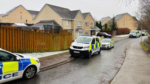 Police cars and vans with blue and yellow checks on a tree-lined street with houses and wooden fencing, and a grey sky.