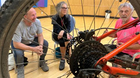 Two men and a woman in a room in a leisure centre, seen through the spokes of an upturned bicycle