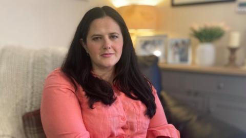 Claire Sugden has long dark hair and is wearing a pink blouse. She is sitting on a light-coloured chair and staring at the camera. In the background is a grey sideboard with a light wooden top. There are framed photographs, a vase of flowers and a brown candlestick with a white candle on top of it.