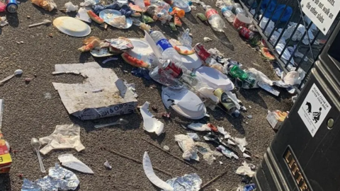 A close-up of overflowing rubbish from a bin strewn across a promenade, next to a black bin and black railings.