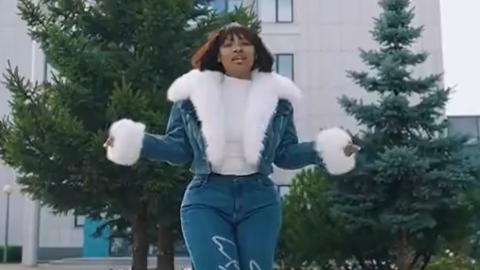 Cyan Boujee wearing jeans and a fur-lined jean jacket stands in front of two cedar trees and an office building. She is holding her arms out as she addresses the camera.
