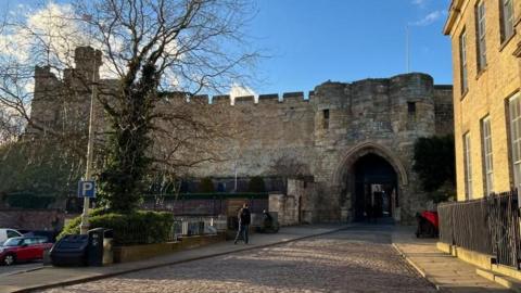 General view of the outside of Lincoln Castle, with a car park and street leading up to the arched entrance.