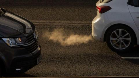 Two cars, one black and one white, driving down a road. There are fumes coming out of the exhaust of the white car.