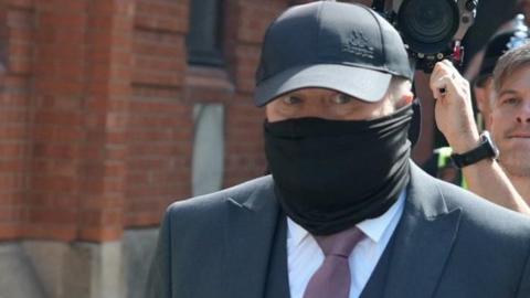 Robert Bush arrives at Hull Crown Court. He is wearing a dark suit and purple tie, a dark-grey baseball cap and a black fabric scarf/wrap, which covers much of his face below his eyes. In the background, is a man is holding up a camera and recording the scene.