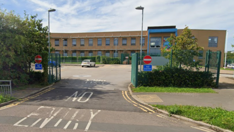The image shows the entrance to a car park in front of a school. The building has a light brown exterior with rectangular windows in a row along the upper floor. In the foreground, the road surface has large white painted text reading no entry. On both sides of the entrance, there are red no entry signs mounted on green metal gates or posts. 