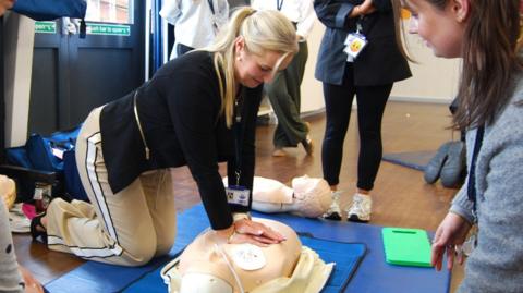 A woman, with long blonde hair, tied back, wearing a black top and cream trousers, leaning over a dummy trying out resuscitation skills. There is another woman wearing and grey top to the right and other people standing around, but you can only see their legs. They are in a  school room with a double door to the left. 