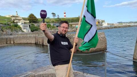 A man posing holding a BBC Radio Devon purple microphone in his right hand, and a large green, black and white Devon flag in his left hand. Behind him is Plymouth's coast line, where you can see Smeaton's Tower. 