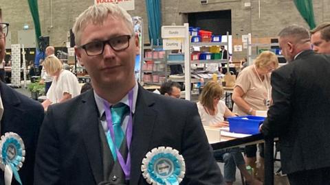 Darren Rance at an election count. He is wearing a suit and tie, black-framed glasses and a blue and white Reform UK rosette. Election staff are counting votes on tables behind him.