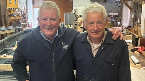 Two men in overalls stand next to the vessel they have been painting and sanding
