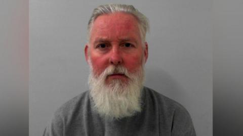 The image shows a man with silver grey swept back and a long, grey beard. He is wearing a plain, dark crew‑neck top and is sitting in front of a grey background, facing the camera.