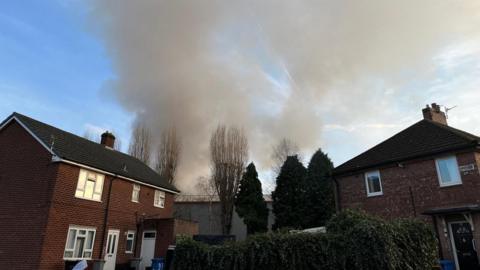 Grey plumes rise into the sky above two red brick houses. The houses are separated by a garden and some trees.