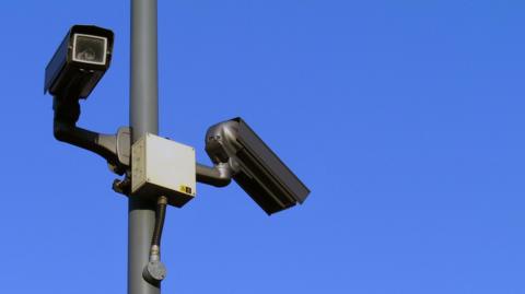 Two CCTV cameras, one pointing towards the viewer and one pointing away. They are set against the backdrop of a vivd blue sky.