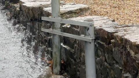 The stolen road sign. It is a metal sign with two poles coming out of the ground. Behind the sign is a rock wall.