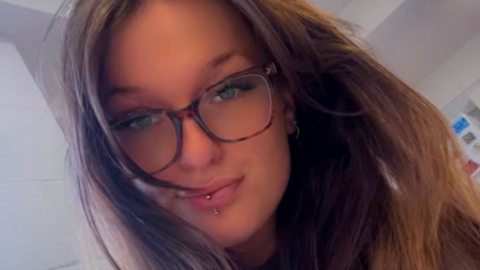 A selfie of a woman with long brown hair and brown glasses. She has a piercing through her bottom lip