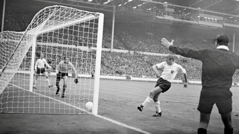 A man in an England top tapping a white football into a goal. Huge crowds watch on. It's a black and white image.