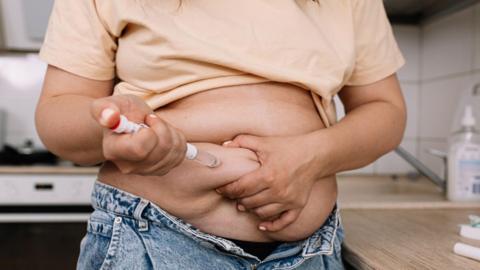 A woman uses a pre-filled injection pen to administer a dose. She is lifting her t-shirt to be able to inject.
