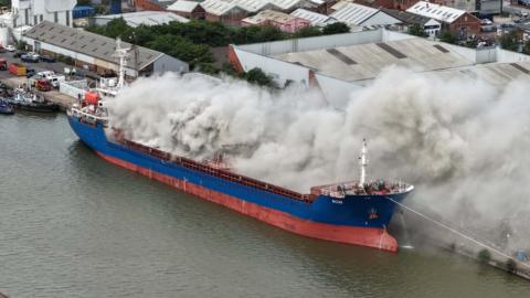 A large plume of grey smoke envelops a large cargo ship. It is visible behind a set of lock gates.