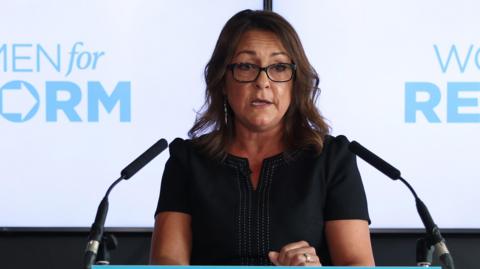 Linden Kemkaran making a speech behind a podium. She is wearing black glasses and a black dress.