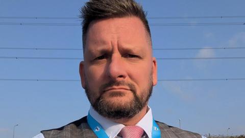 Joel Tetlow has short brown hair and a beard. He is wearing a shirt, waistcoat and pink tie, with the top of a Reform UK lanyard just visible.