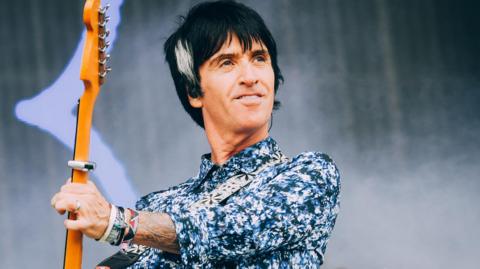 Johnny Marr, with black hair and wearing a blue, white and black speckled shirt, holds the neck of an orange guitar aloft