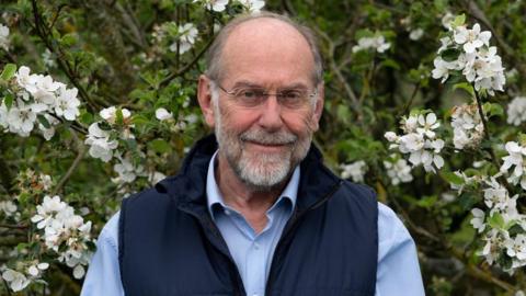 A man with receded grey hair and a beard and glasses stands among white flowering branches. He is wearing a blue gilet over a light blue shirt