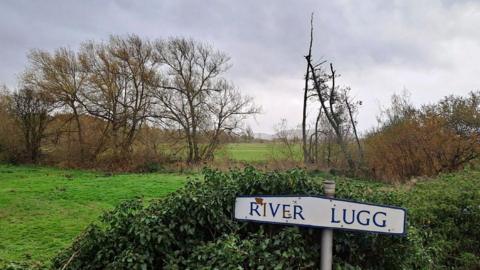 In the foreground is a sign which reads "River Lugg" while in the background is a meadow with trees and bushes.