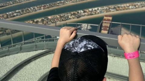 From behind, you see a young boy with a black baseball cap has his hands on a silver railing looking out from a height onto strips of land within water, heavily populated with buildings. He has a pink tourist wristband on his right arm.