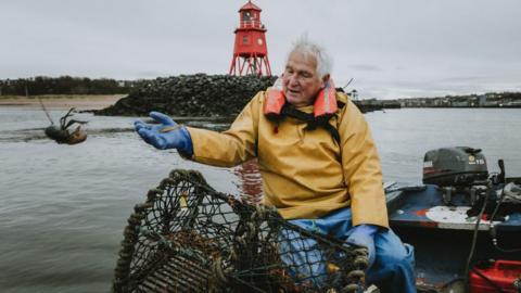 A white-haired man in yellow water proof clothing is sitting on a boat and throwing a lobster back into the sea. He is not far from the shore.