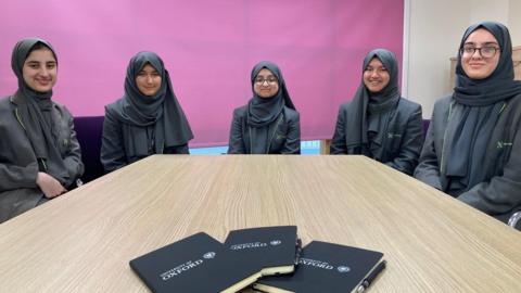 Five schoolgirls wearing black headscarves sitting at a table. 