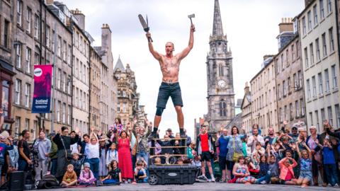 A street performer holding several sharp objects including a knife and an axe in his hands while he balances on stacked wooden boards in front of a large crowd of peoiple and several old light-stone buildings on the Royal Mile in Edinburgh.