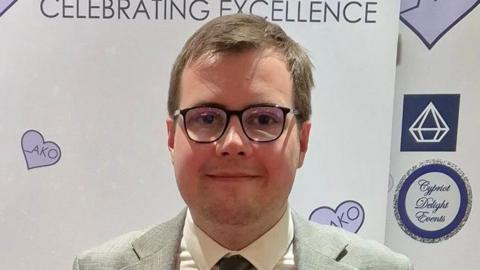 A head and shoulders image of Tom Howard, who is wearing glasses, a grey suit, white shirt and dark grey tie. He is looking straight into the camera. 