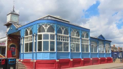 Blue fronted wooden cafe with intricate ironwork decorative framework. The bottom of the cafe is painted red. There are 2 bins standing to the left of the cafe which is near a road and there are steps either side. Behind the cafe building is an ornate Victorian clock tower.