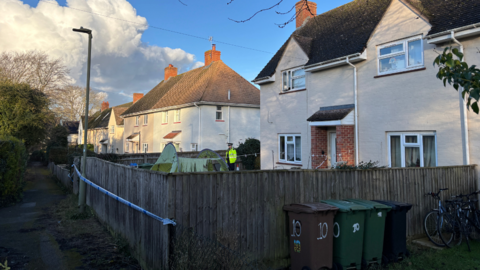 A police officer stands next to a white house, which is surrounded by police tape.
